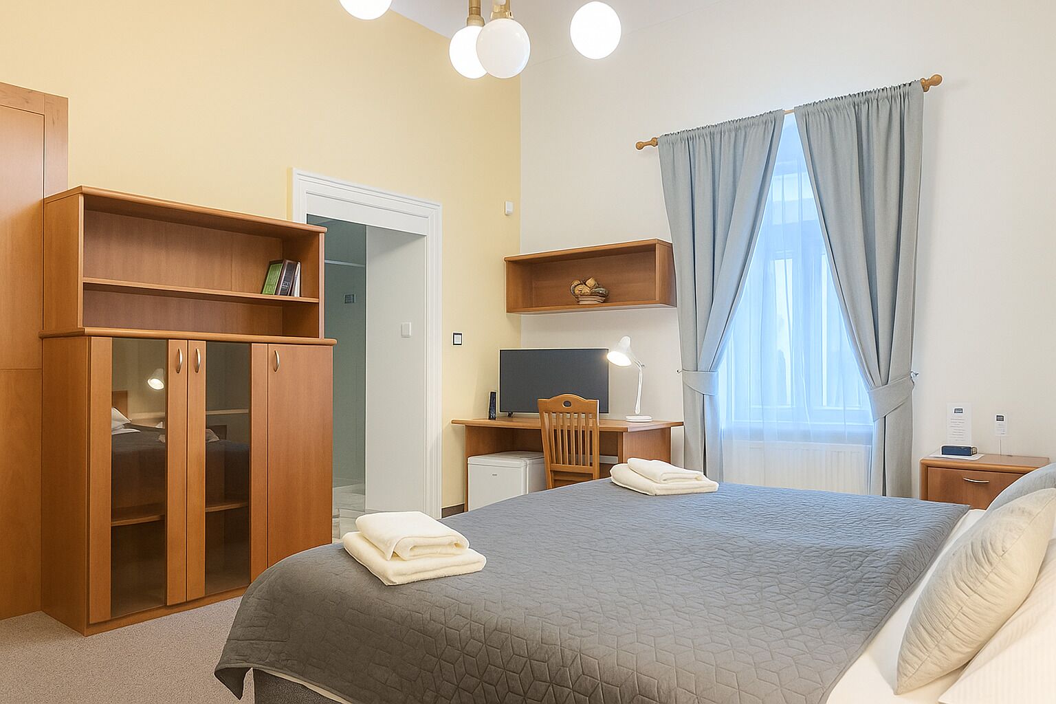 A hotel room with a bed, a desk, a computer, a wardrobe, and a window with curtains.