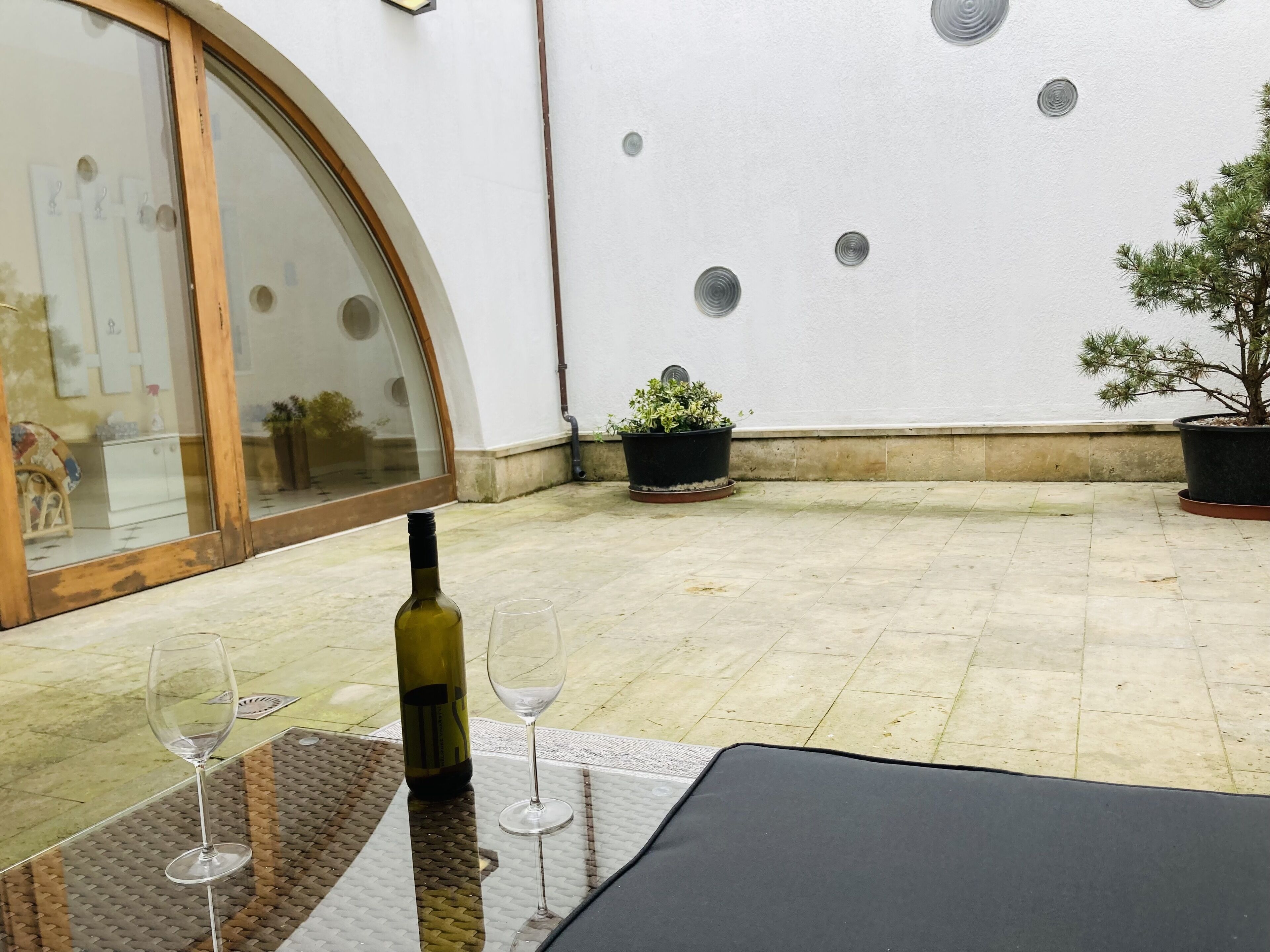 Een terras met een tafel, een fles wijn en glazen, een bloempot en een muur met ronde patronen.