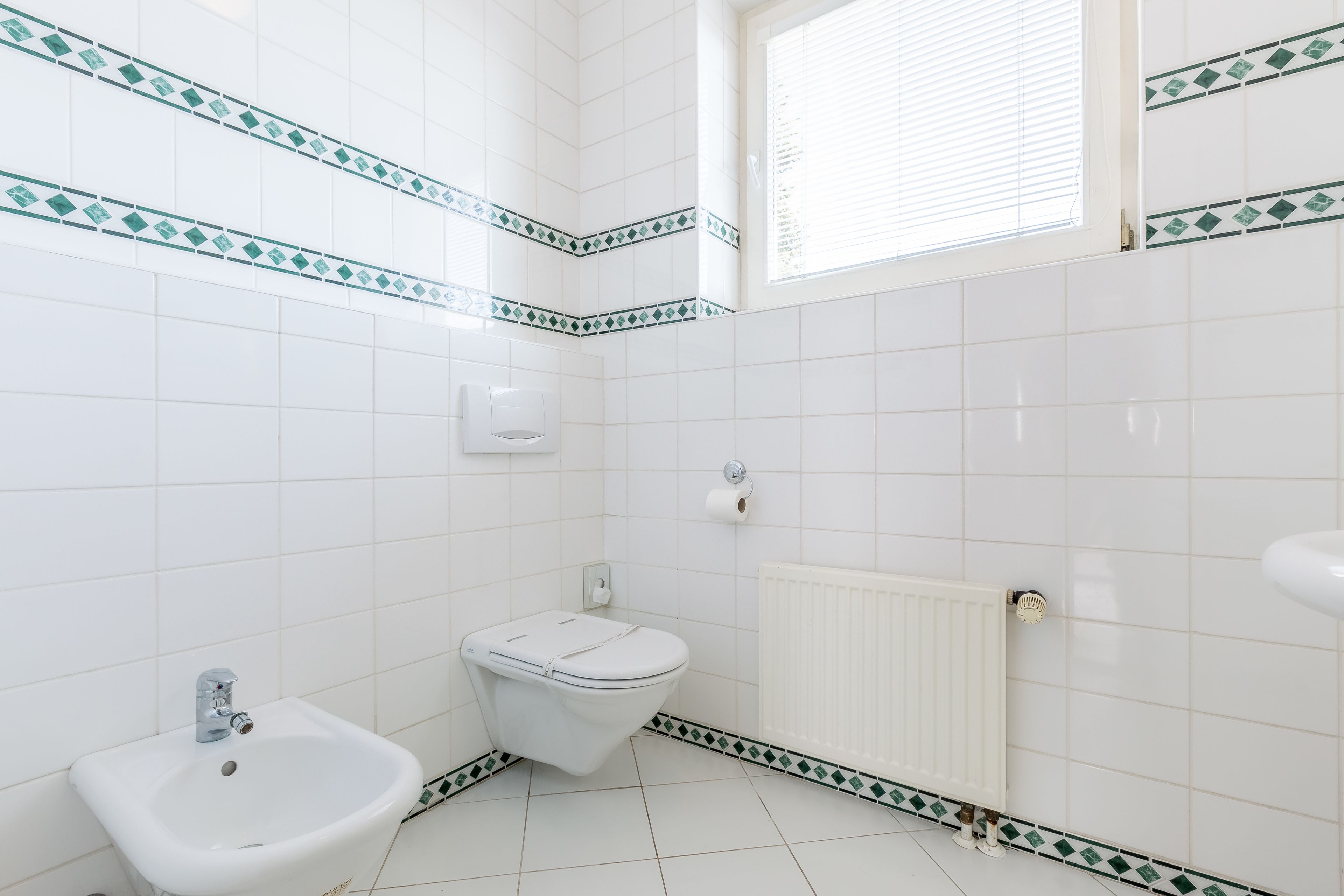 Un baño con azulejos blancos, un bidé, un inodoro, un radiador y una ventana con persianas.