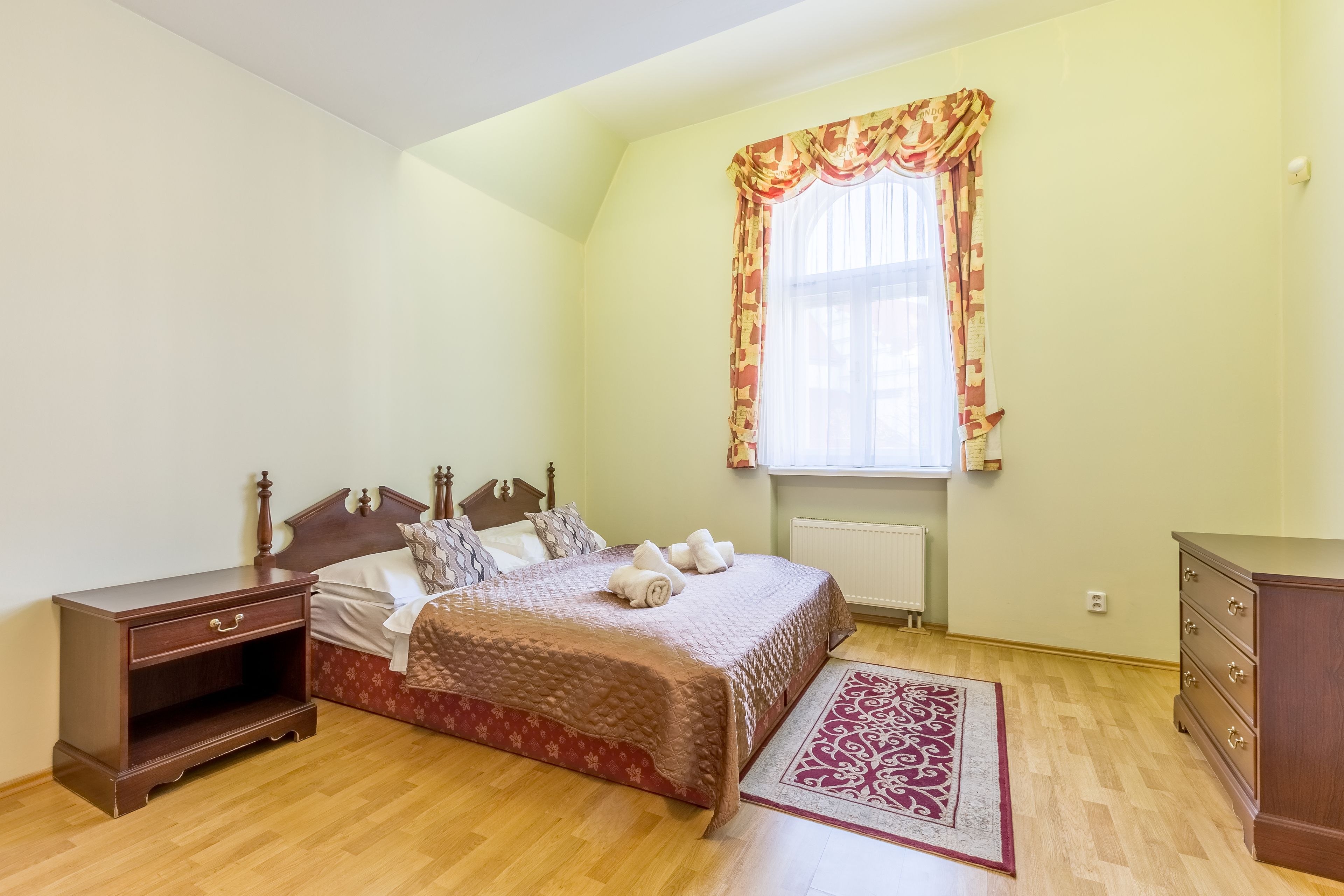 A bedroom with a wooden bed, a nightstand, a dresser, and a window with curtains.