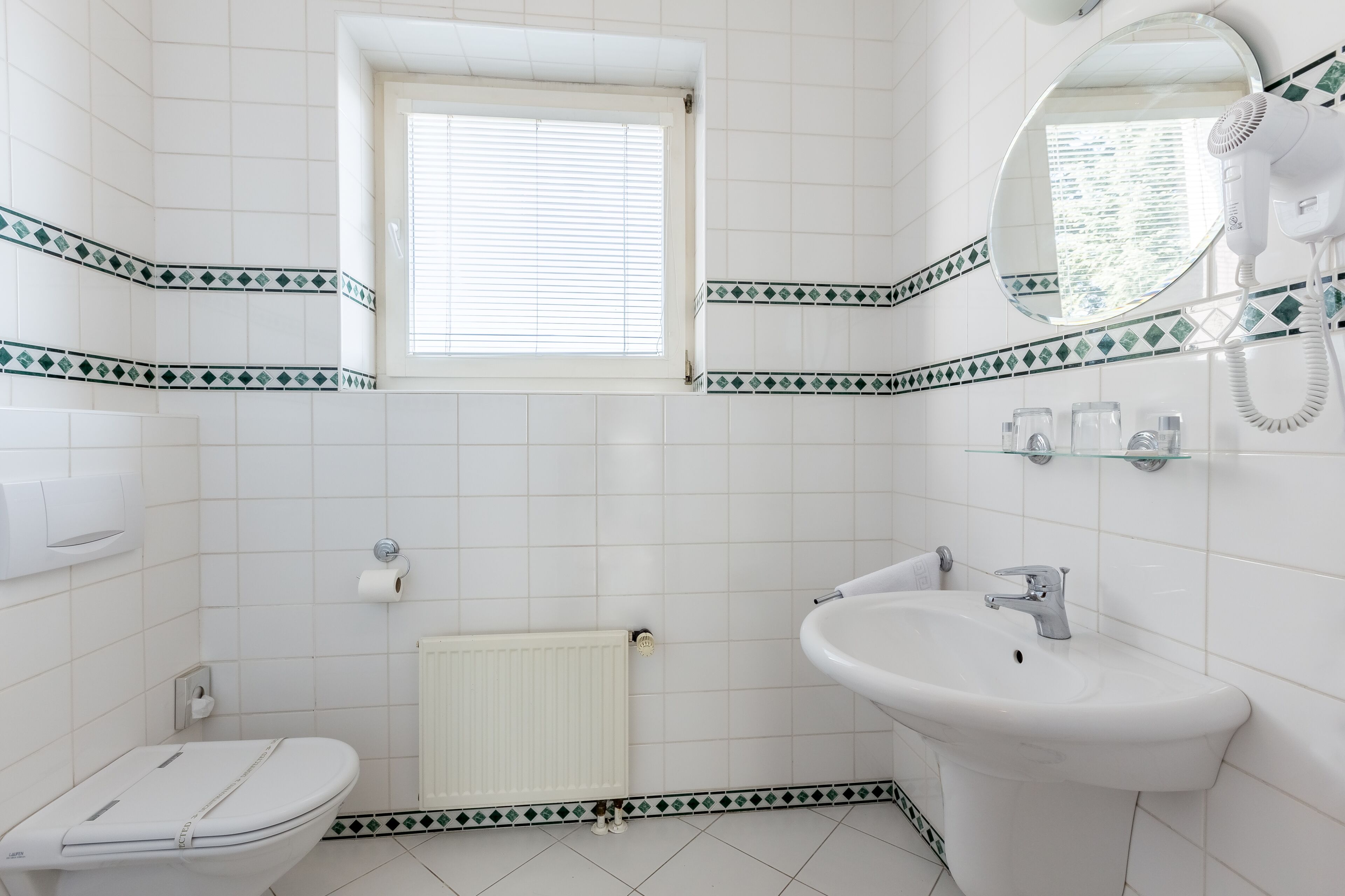 A bathroom with white tiles, a toilet, a sink, a mirror, and a hairdryer.
