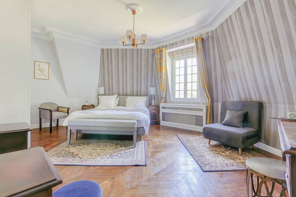 Een slaapkamer met een bed, twee stoelen, een bureau, een radiator en een raam met gordijnen.