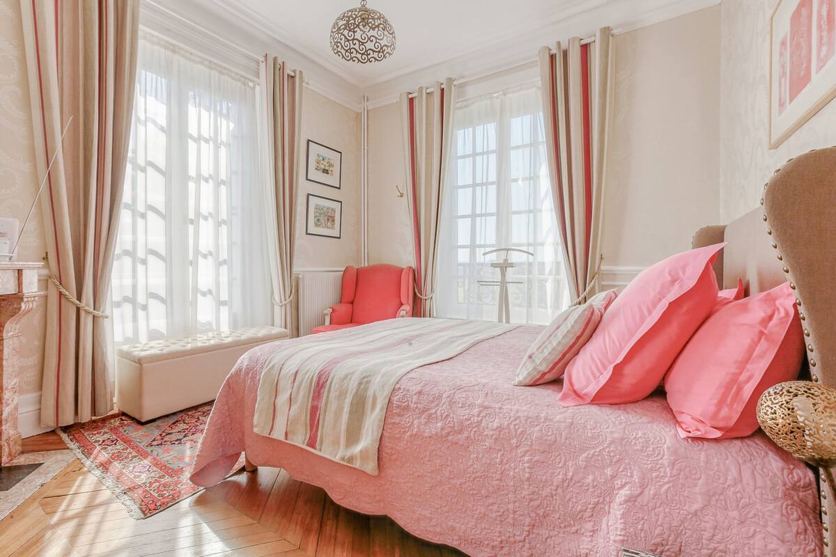 Een slaapkamer met een groot bed, roze beddengoed, een houten vloer en een raam met gordijnen.