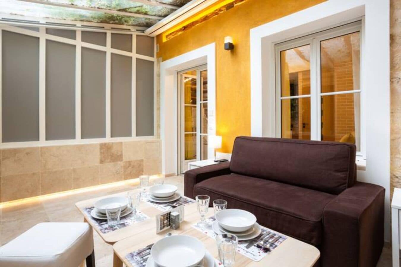 A dining area with a brown sofa, a table set with plates and glasses, and a window with a view of a brick building.