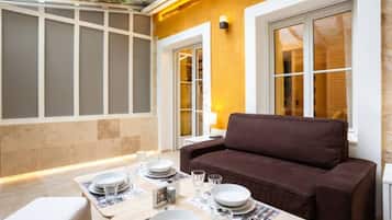 A dining area with a brown sofa, a table set with plates and glasses, and a window with a view of a brick building.