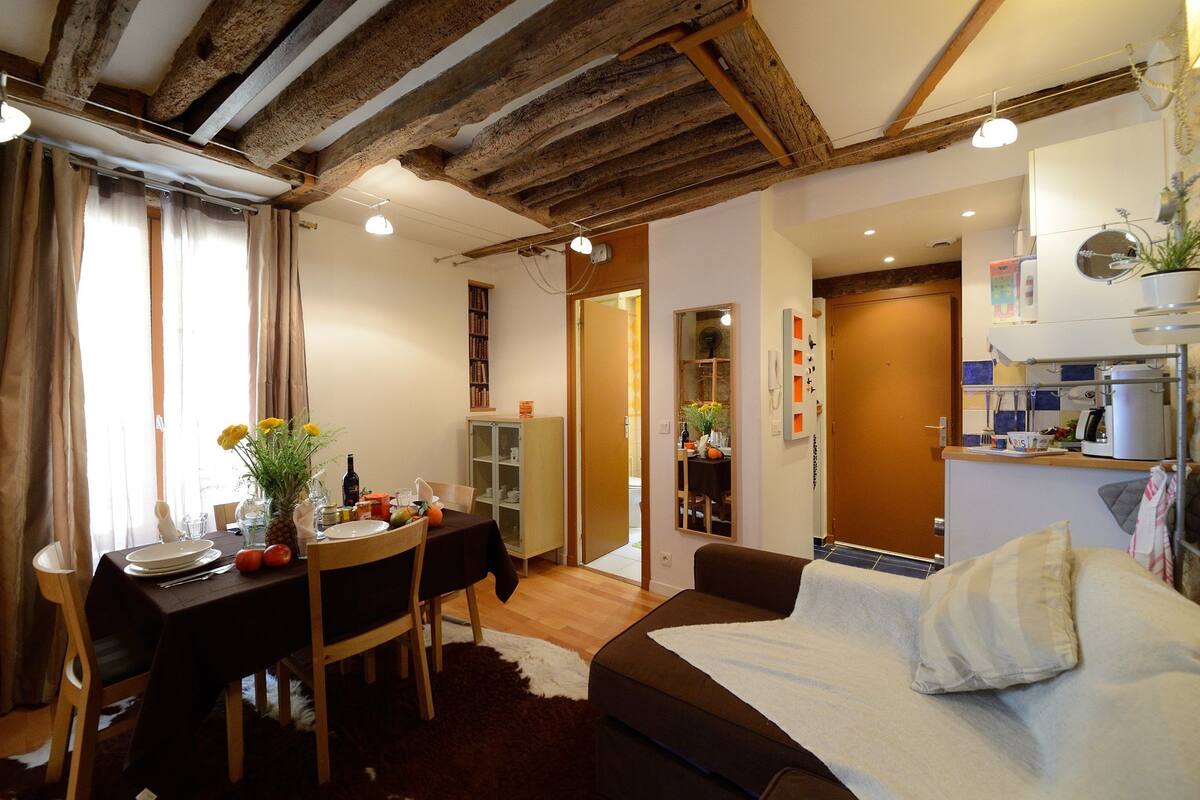 A cozy room with a wooden ceiling, a dining table set with plates and a vase of flowers, a sofa with a blanket, and a kitchen area with a coffee machine.