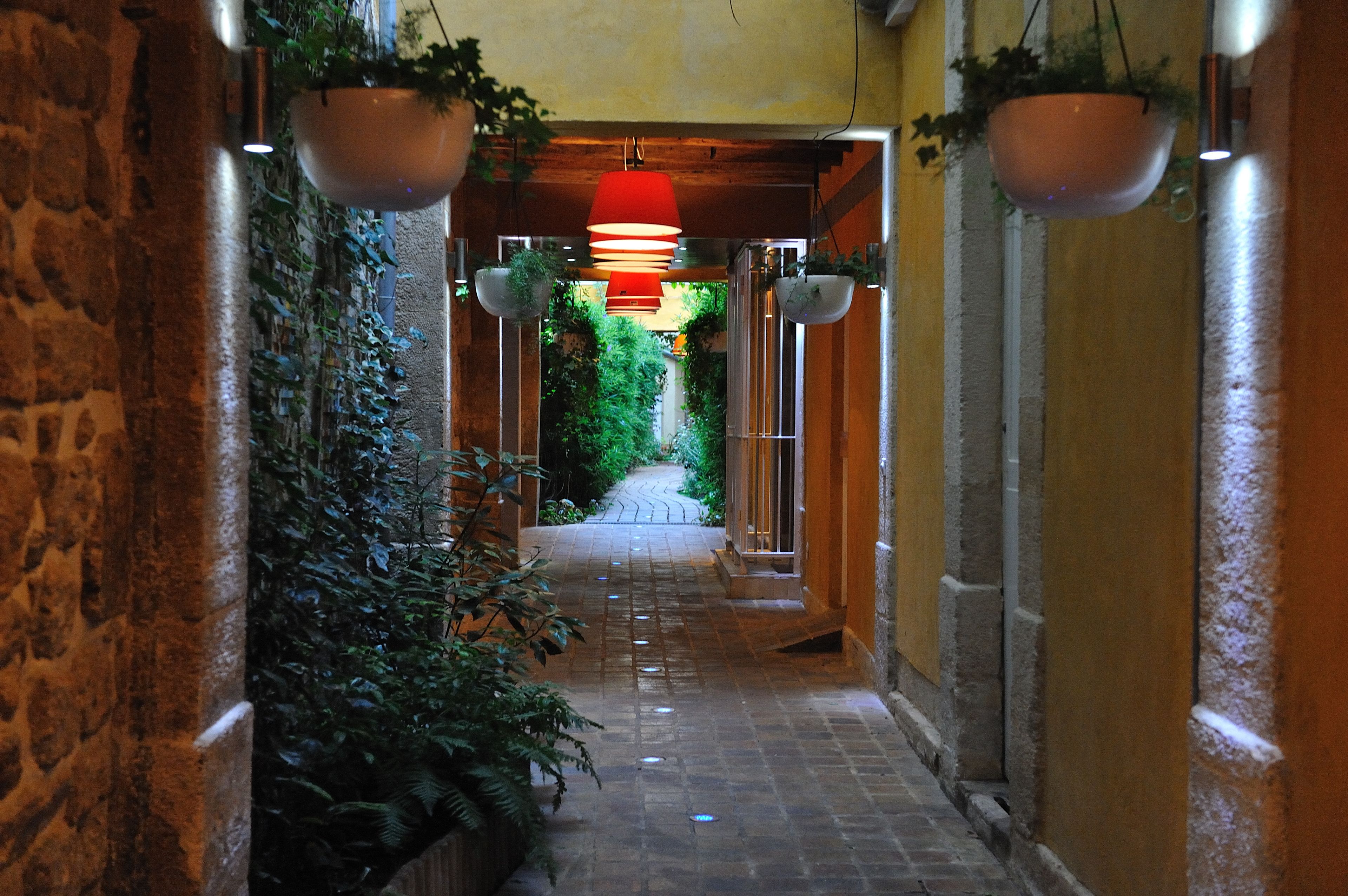 Un corridoio stretto con pareti in pietra, fioriere pensili con piante verdi e un lampadario rosso.