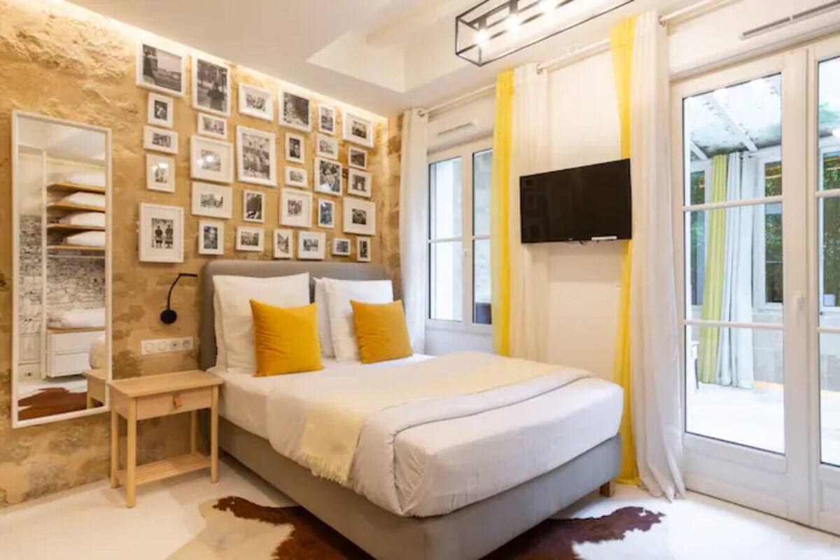 A bedroom with a bed, a bedside table, a wall-mounted TV, and a large window with curtains.
