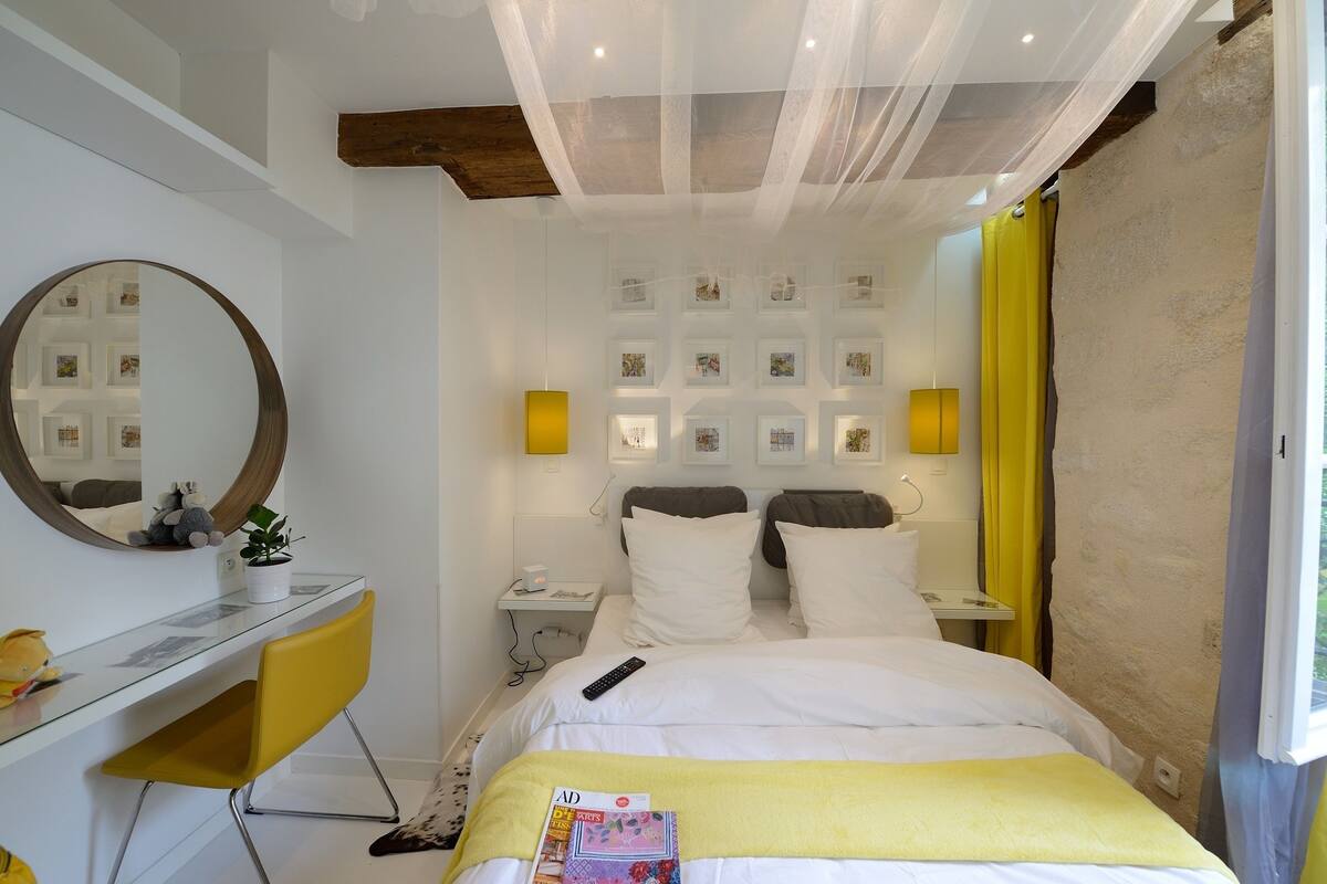 A hotel room with a bed, a desk with a chair, a mirror, a bedside table, and a wall with framed pictures.