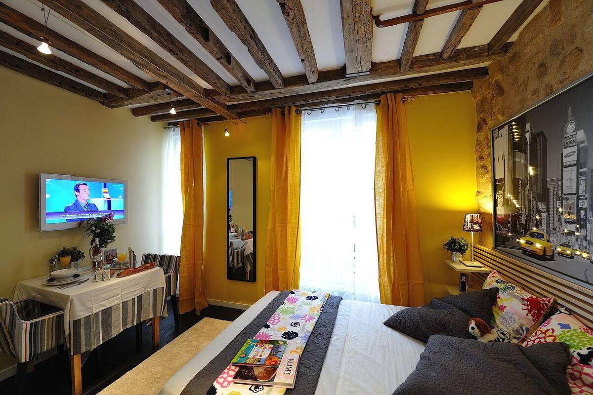 A hotel room with a bed, a dining table with chairs, a TV, and a cityscape painting on the wall.