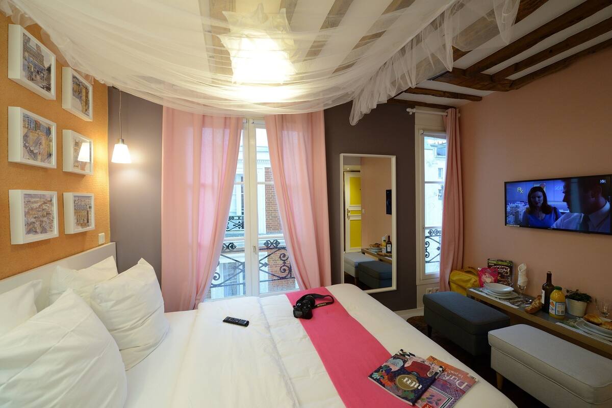 A hotel room with a bed, a TV, a balcony with pink curtains, and a dining area with a table and chairs.