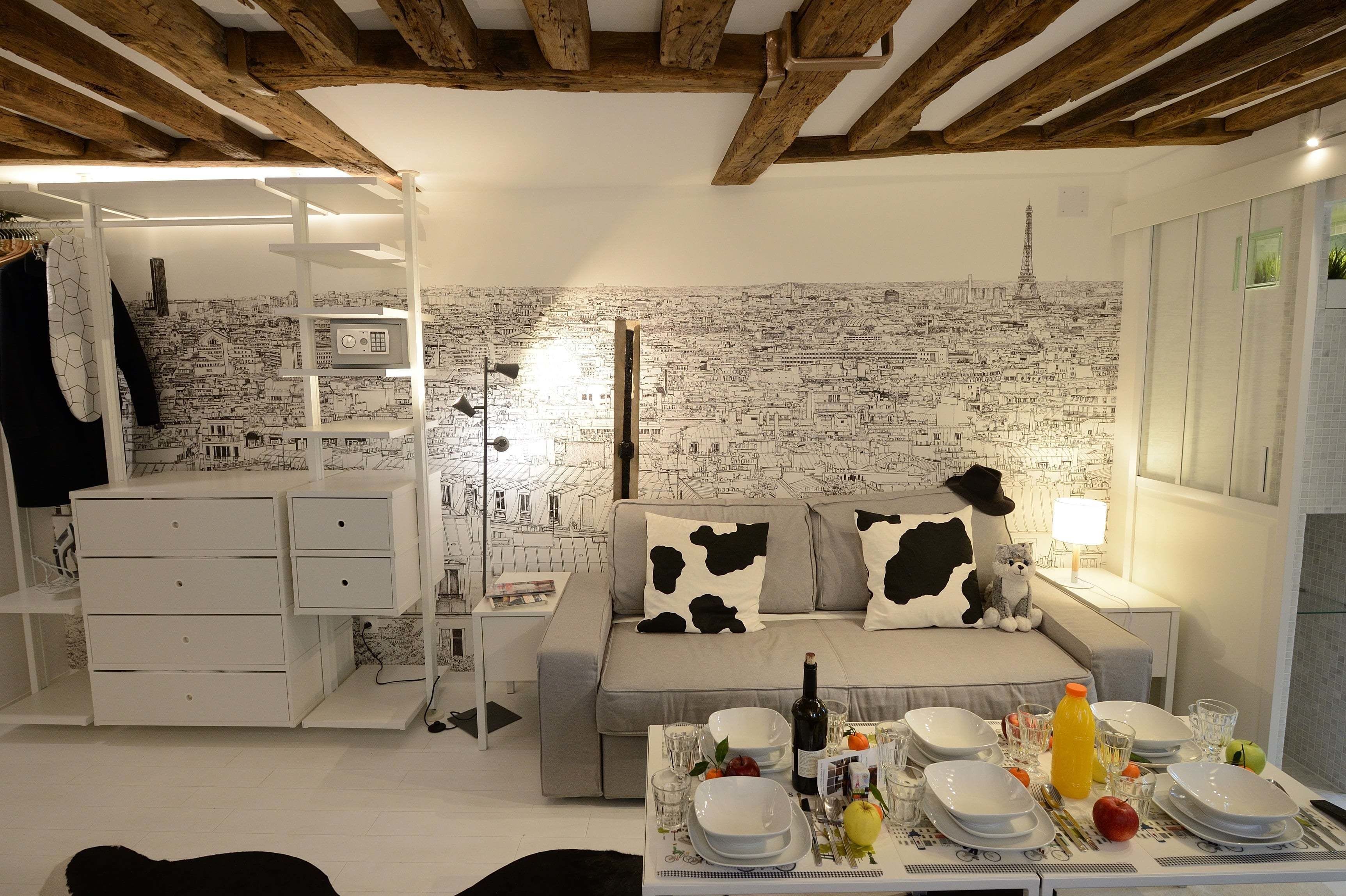 A cozy room with a cityscape mural, a sofa with cow-patterned pillows, a dining table set with plates and glasses, and a white dresser.
