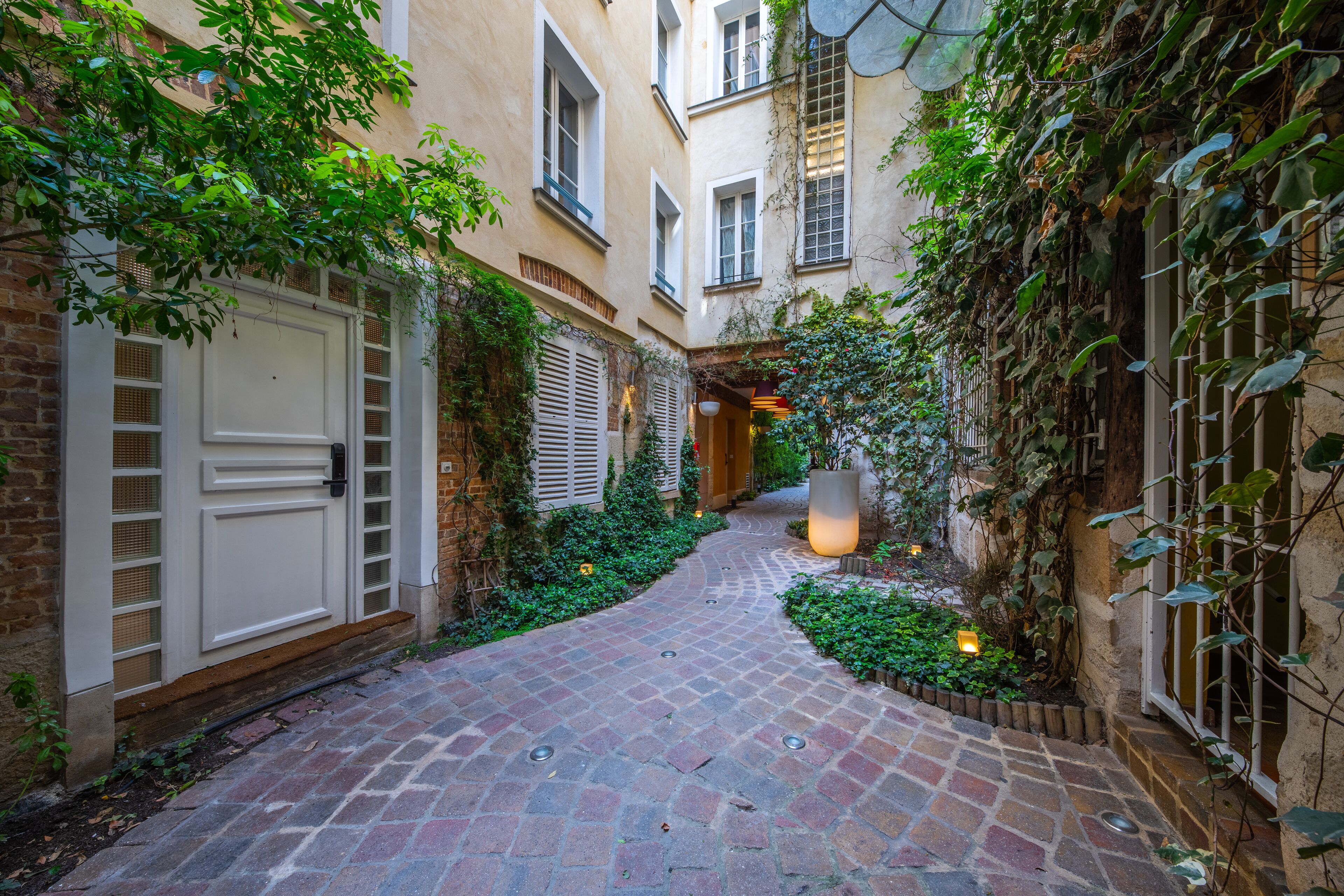 A narrow brick pathway flanked by buildings with white shutters and greenery.
