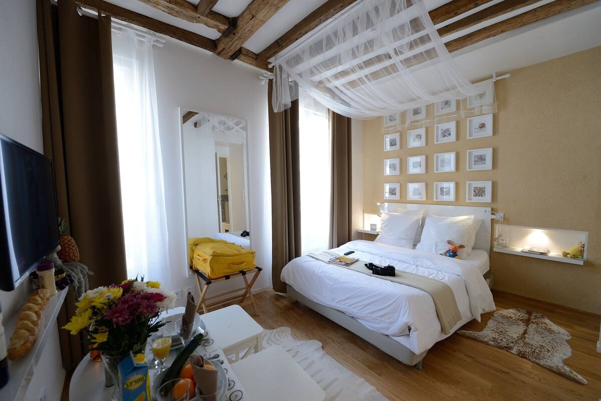 A hotel room with a bed, a TV, a desk with a chair, a mirror, and a wall with framed pictures.