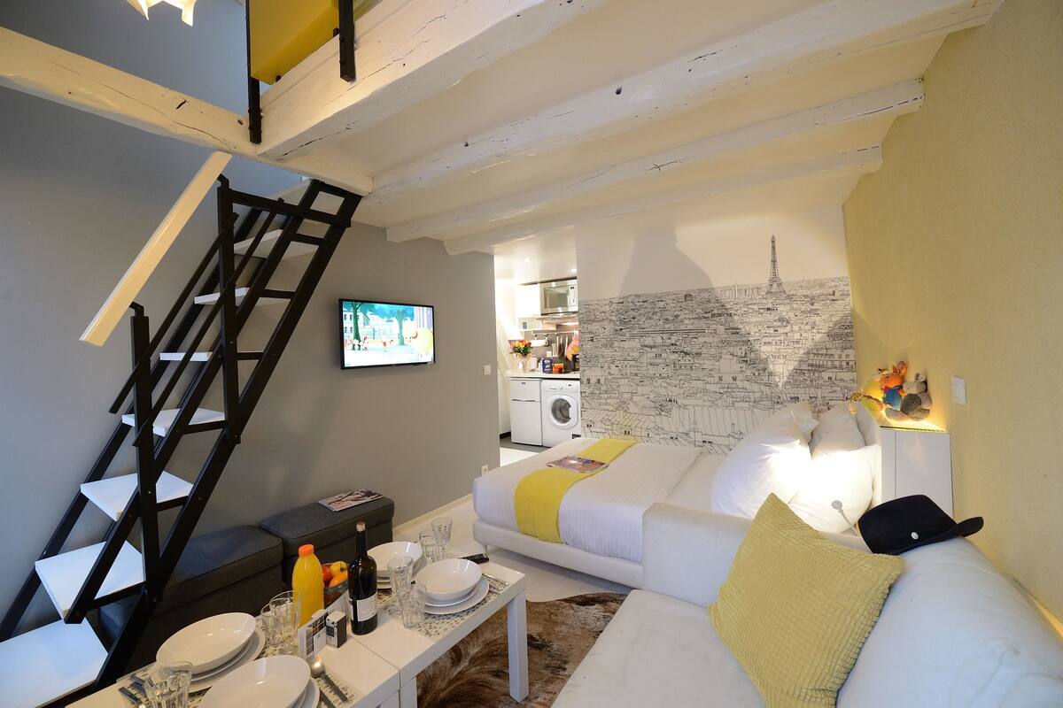 A compact living space with a sofa, a dining table with plates and glasses, a TV, a staircase, and a kitchen area visible through an open door.