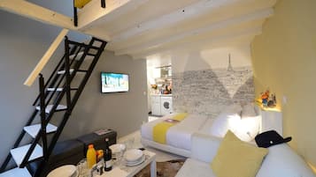 Un ambiente compatto con un divano, un tavolo da pranzo apparecchiato con piatti e bicchieri, una televisione, una scala e una cucina visibile attraverso una porta aperta.