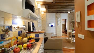 Una cucina con lavello, piano cottura e diversi tipi di frutta sul bancone. Si nota una televisione sullo sfondo e un'area pranzo con tavolo e sedie.