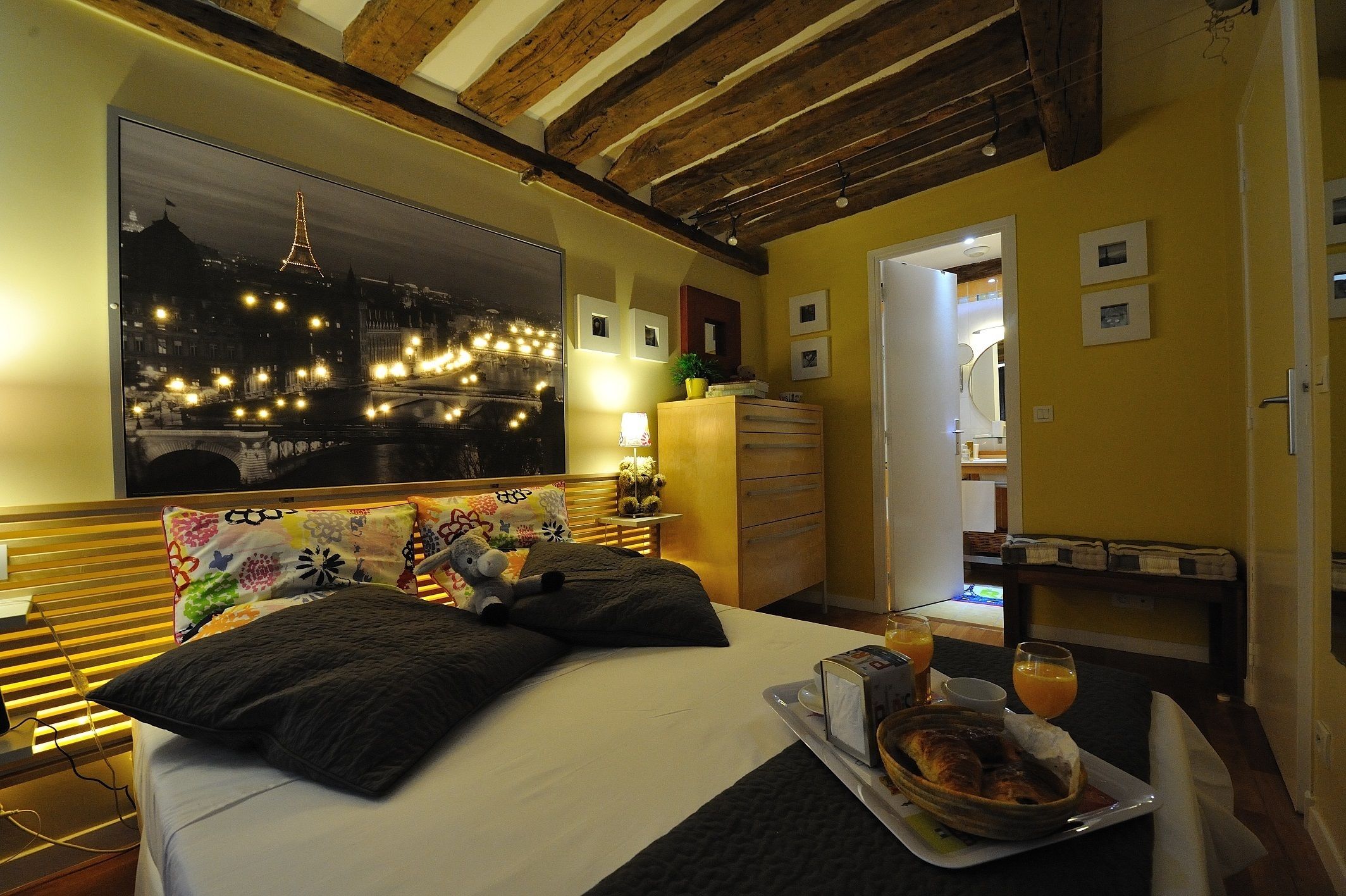 A hotel room with a bed, a tray with breakfast, a night view of a cityscape, and a wooden ceiling.