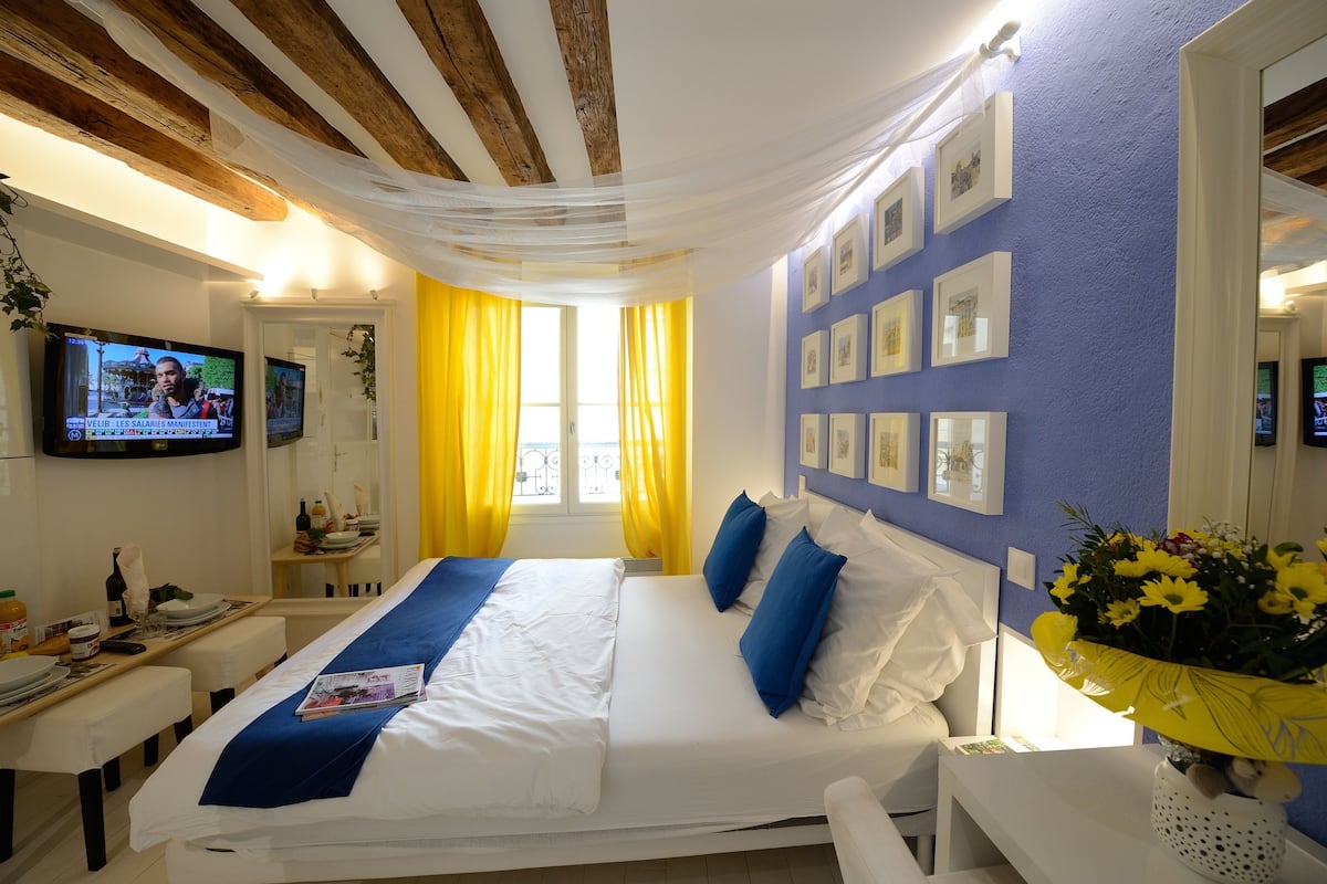A room with a bed, a TV, a window with yellow curtains, and a wall with framed pictures.