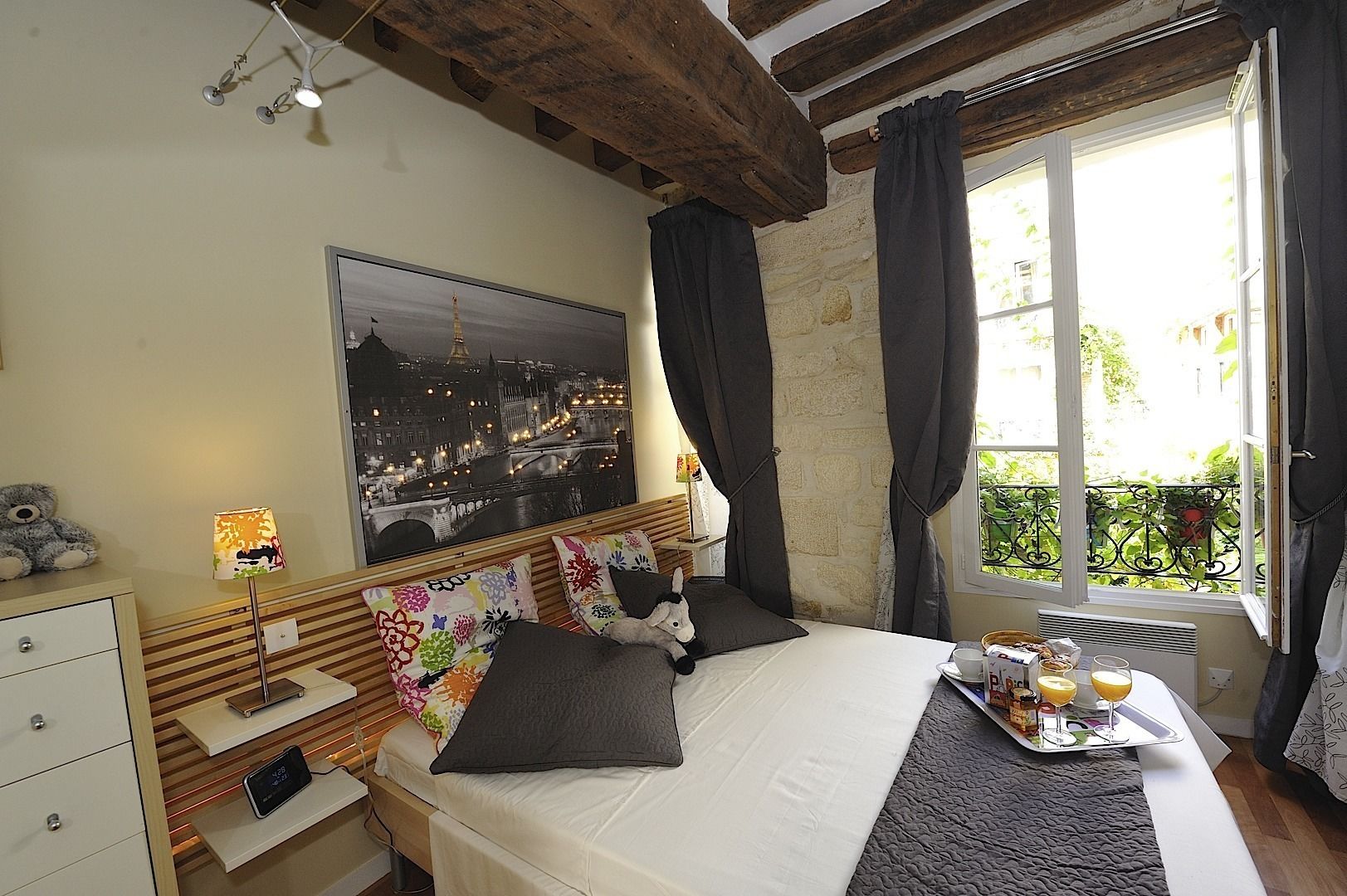 A bedroom with a bed, a nightstand, a lamp, a teddy bear, a tray with breakfast items, and a cityscape painting on the wall.