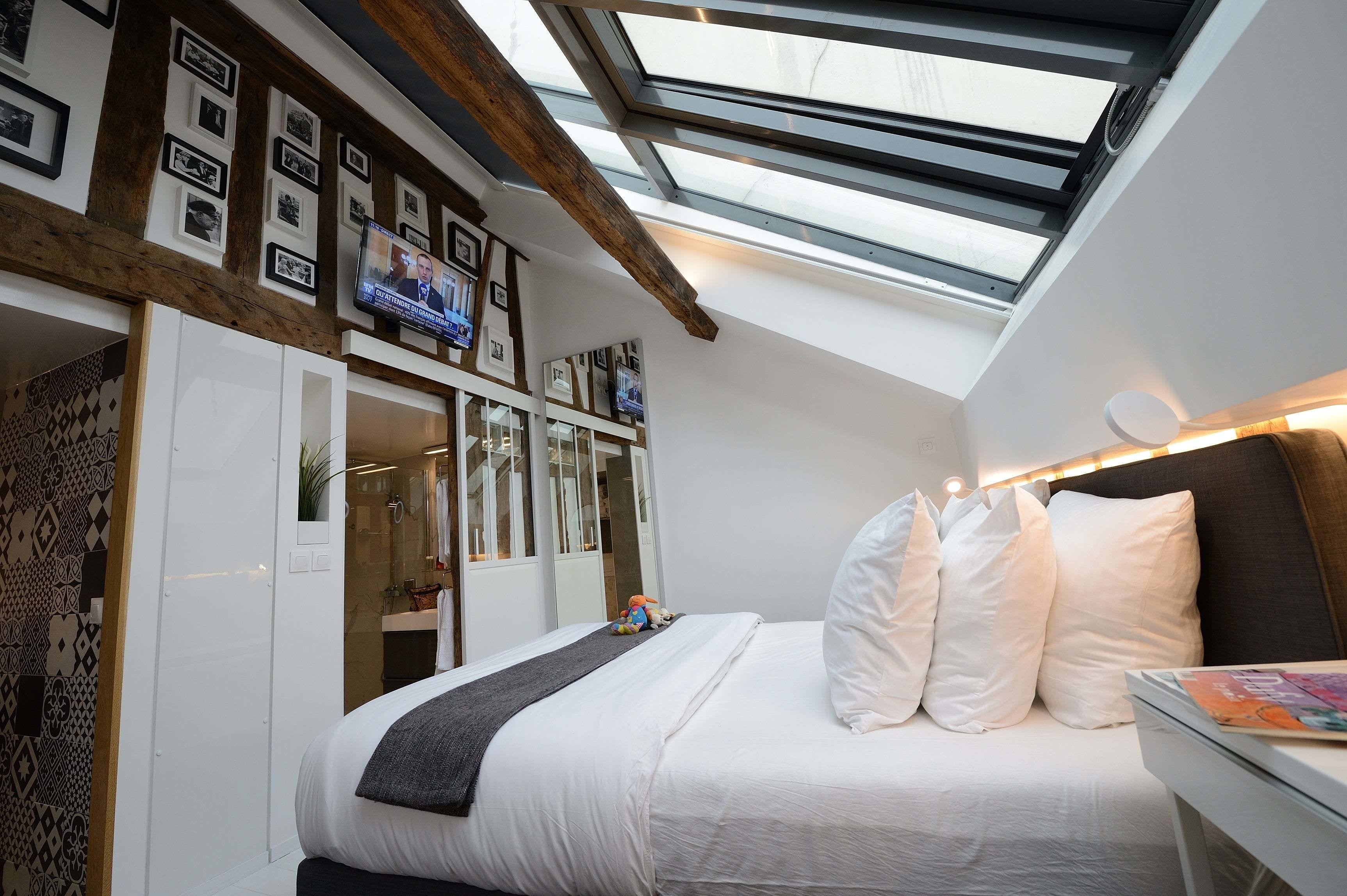 A bedroom with a skylight, a bed with white bedding, a bedside table, and a TV mounted on the wall.