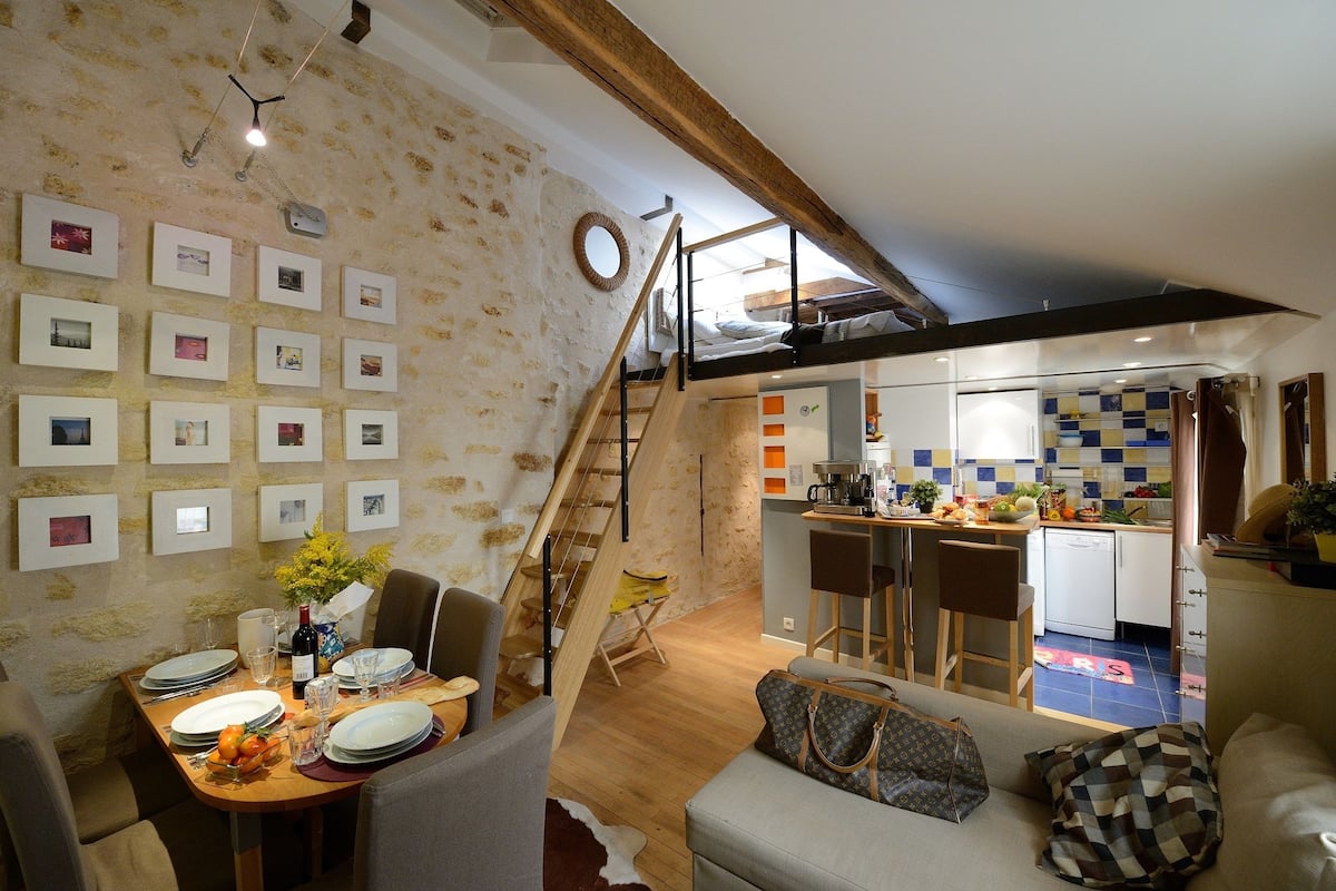 A dining area with a wooden table set for a meal, a staircase leading to a loft, and a kitchen visible through an open door.