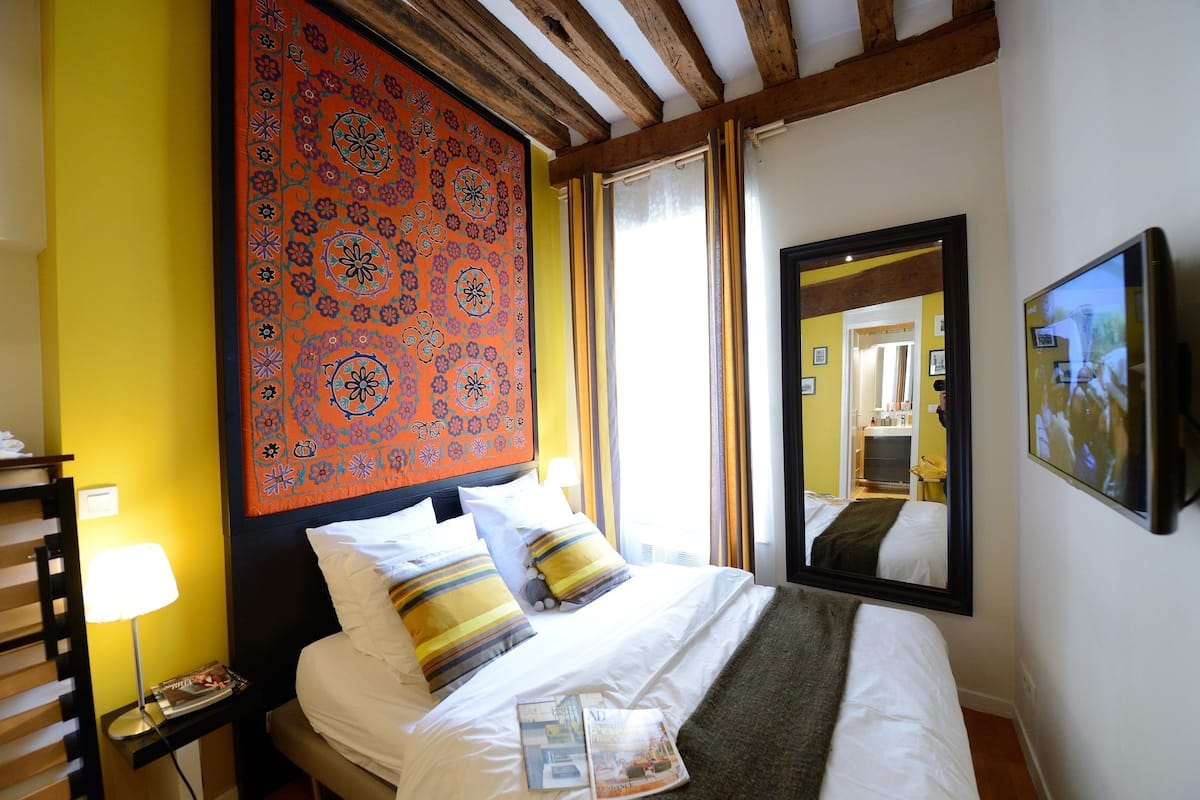 A hotel room with a bed, a nightstand, a lamp, a mirror, and a large tapestry on the wall.