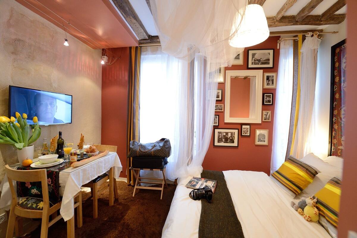 A hotel room with a bed, a dining table with breakfast items, a chair, a suitcase, and a television on the wall.