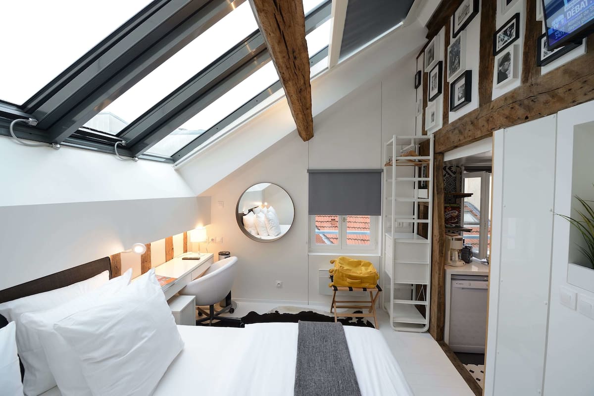 A modern bedroom with a skylight, a bed, a desk, a chair, a mirror, and a small kitchenette.