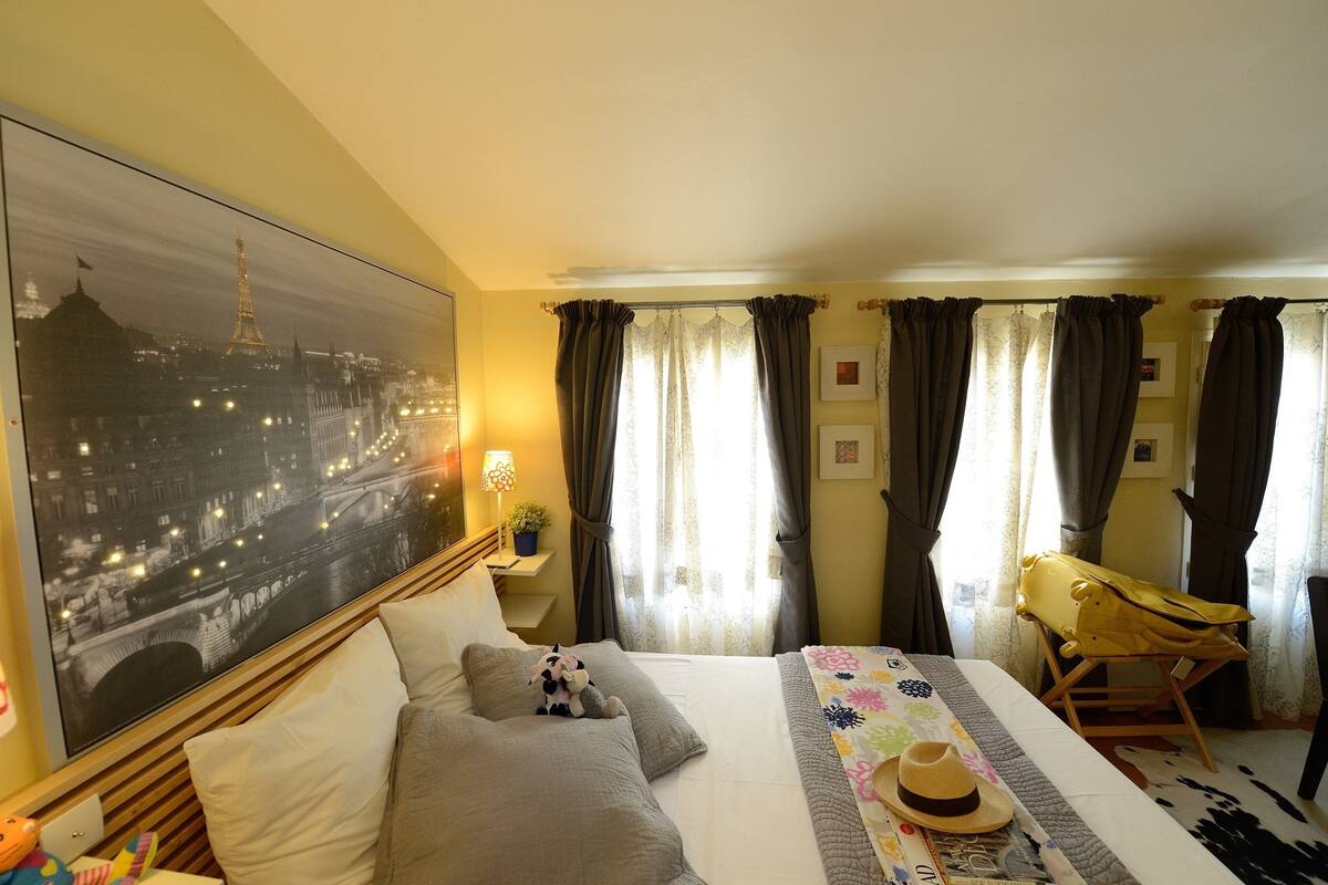 A hotel room with a large bed, a view of the Eiffel Tower, a small table with a lamp, and a chair.