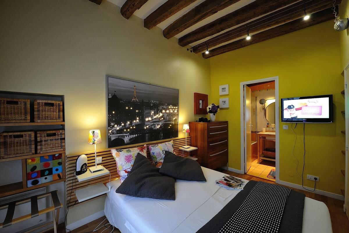 A bedroom with a bed, a TV, a nightstand, a lamp, a wardrobe, and a window with a cityscape view.