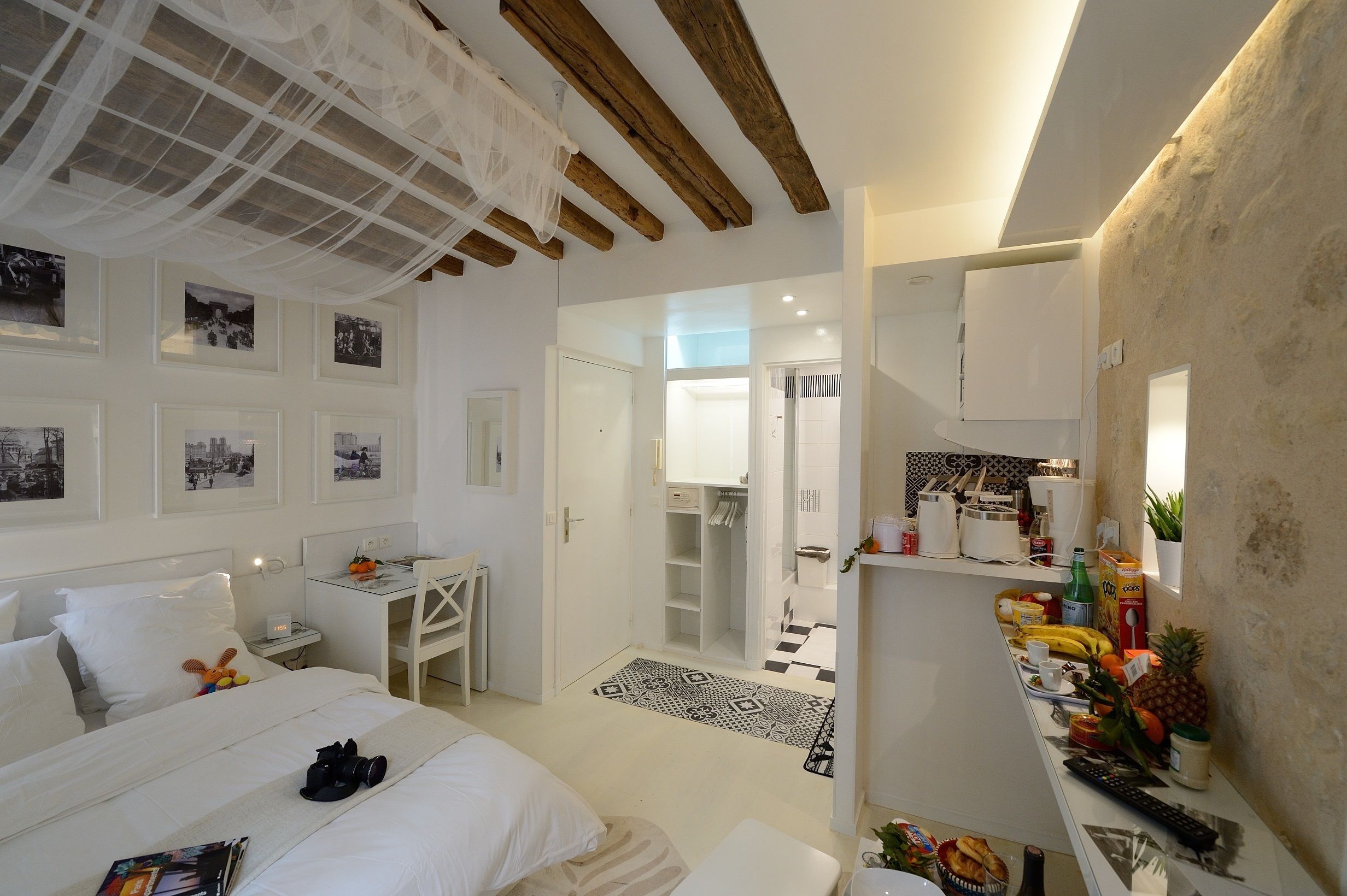 Une chambre d’hôtel avec un lit, un bureau et une kitchenette. On peut voir des tableaux encadrés au mur, une armoire et une salle de bain à travers une porte ouverte.