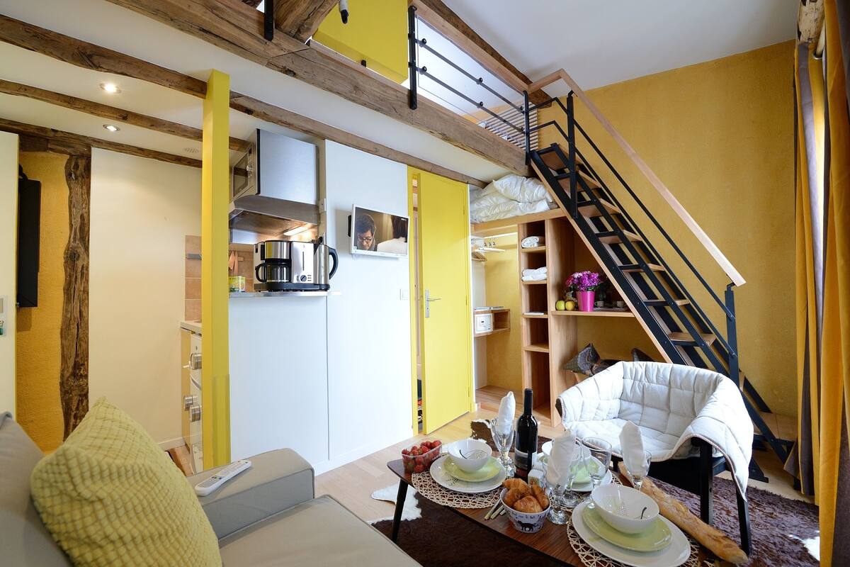 A cozy living room with a sofa, a dining table set with food and drink, a staircase, and a kitchen area.
