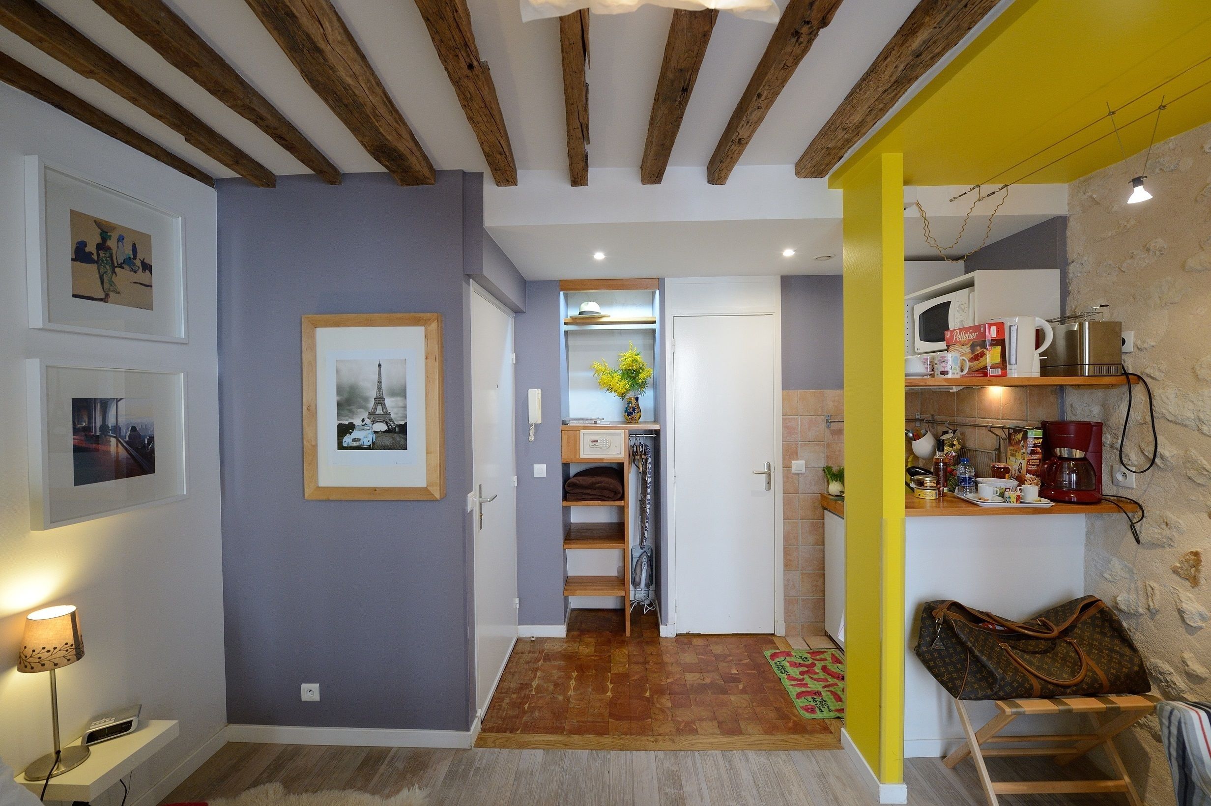 A hallway with a wooden ceiling, a kitchen area with a microwave and coffee maker, and a door leading to a room with a bed.