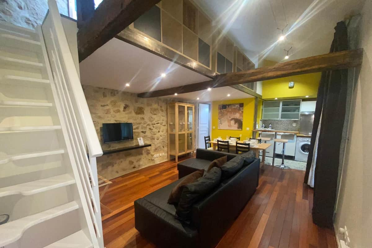 A modern living space with a wooden floor, a black sofa, a stone wall, and a kitchen area.