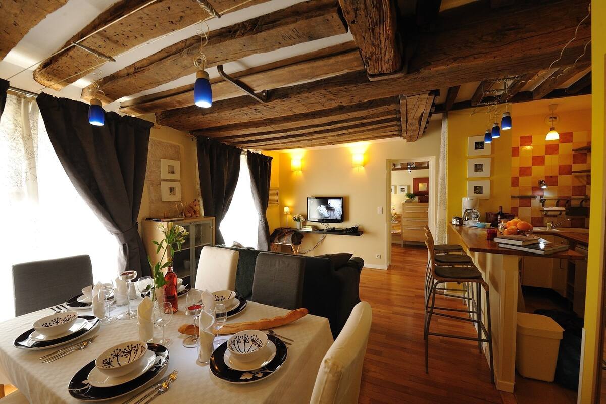 A dining area with a table set for four, a television in the background, and a kitchen area with a bar.