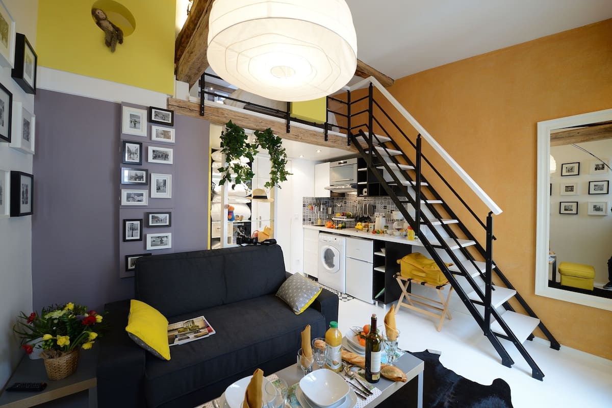 A modern living room with a black sofa, a dining table set with plates and glasses, a staircase, and a kitchen in the background.