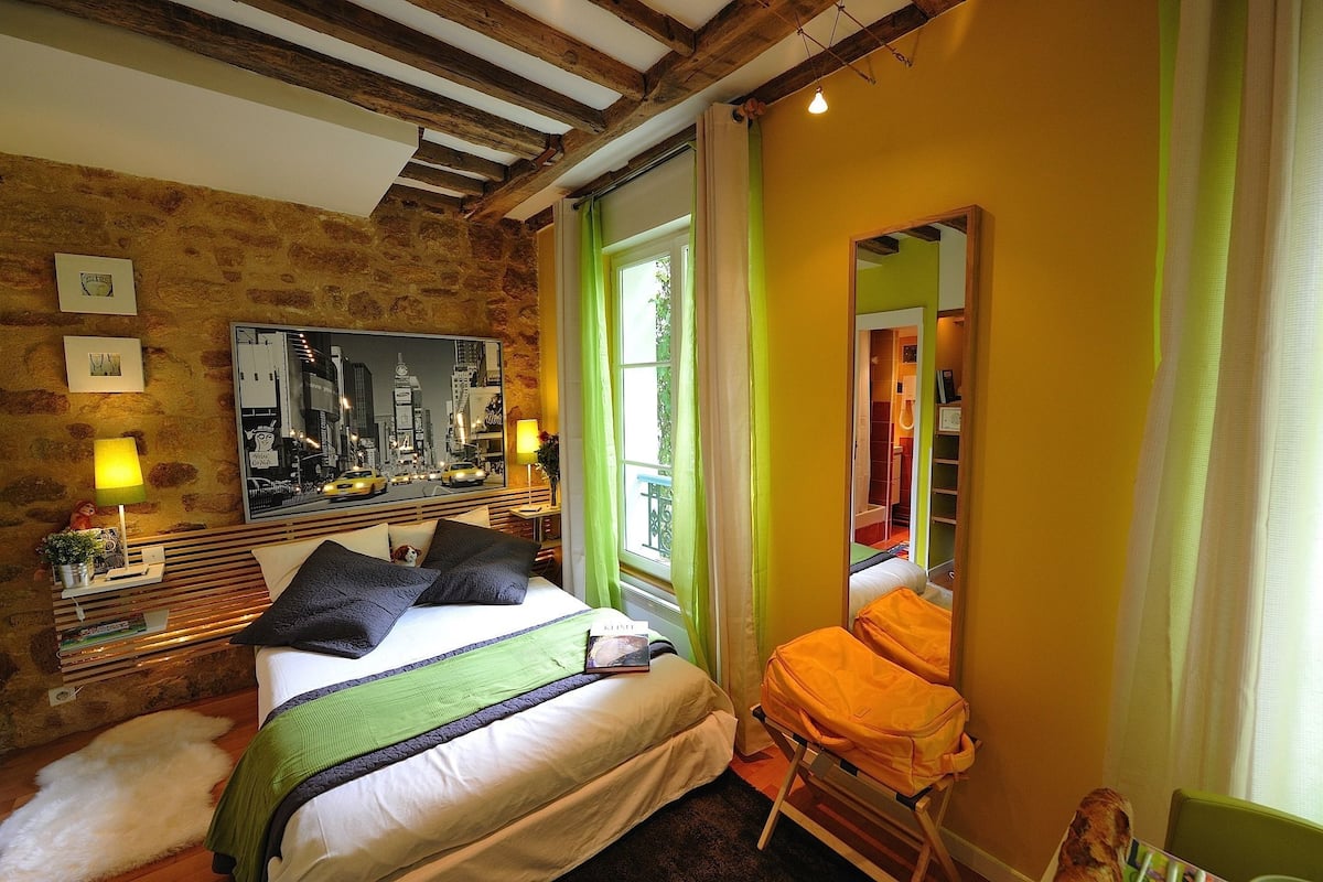 A bedroom with a bed, a mirror, a window with curtains, a wooden headboard, and a cityscape painting.