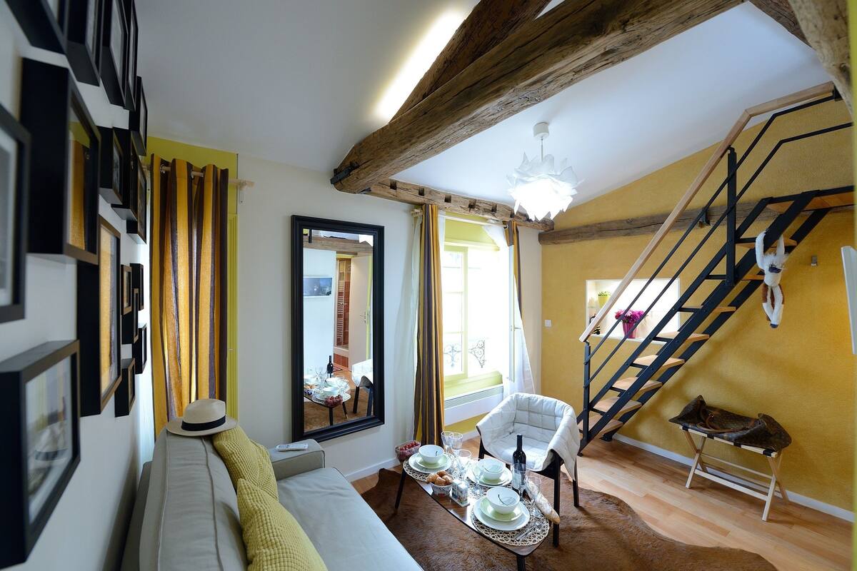 A cozy living room with a sofa, a dining table set for two, a staircase, and a large mirror.