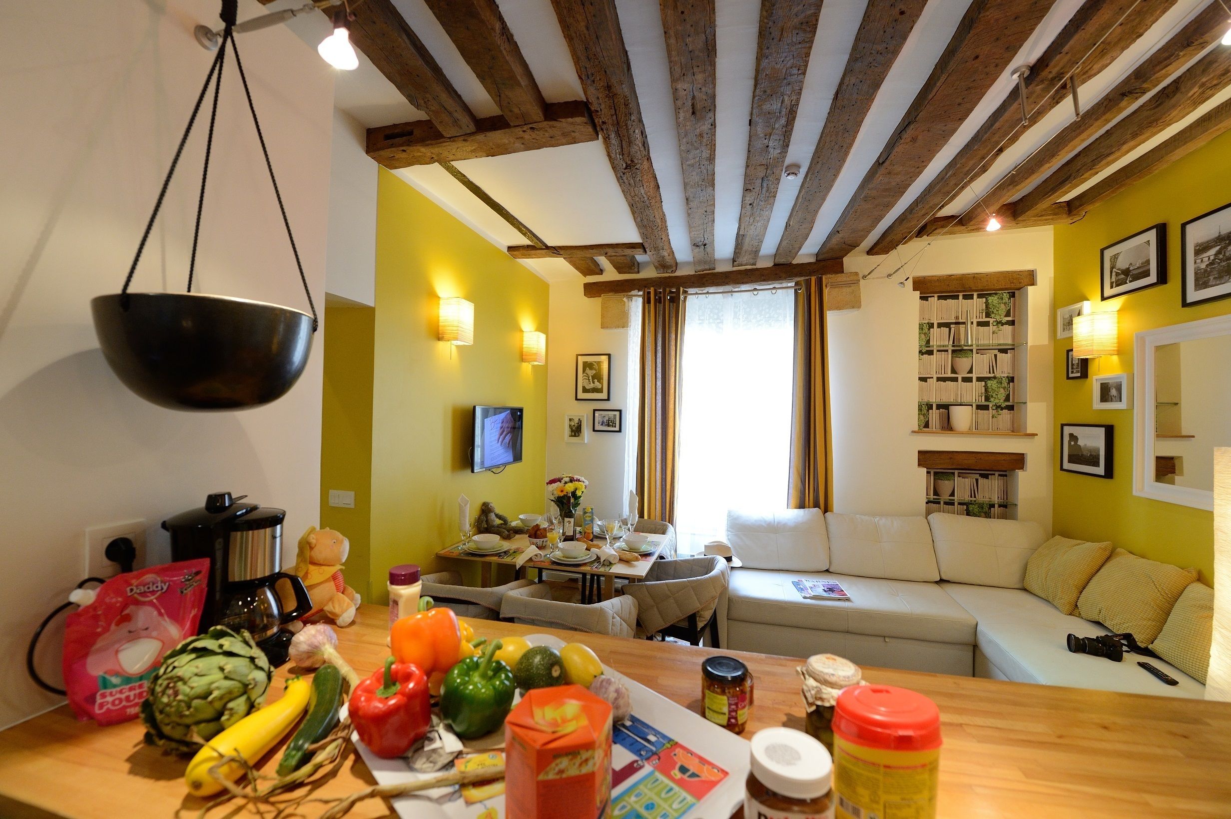 A cozy kitchen with a hanging bowl, a dining table with food, and a sofa in a room with wooden beams and yellow walls.