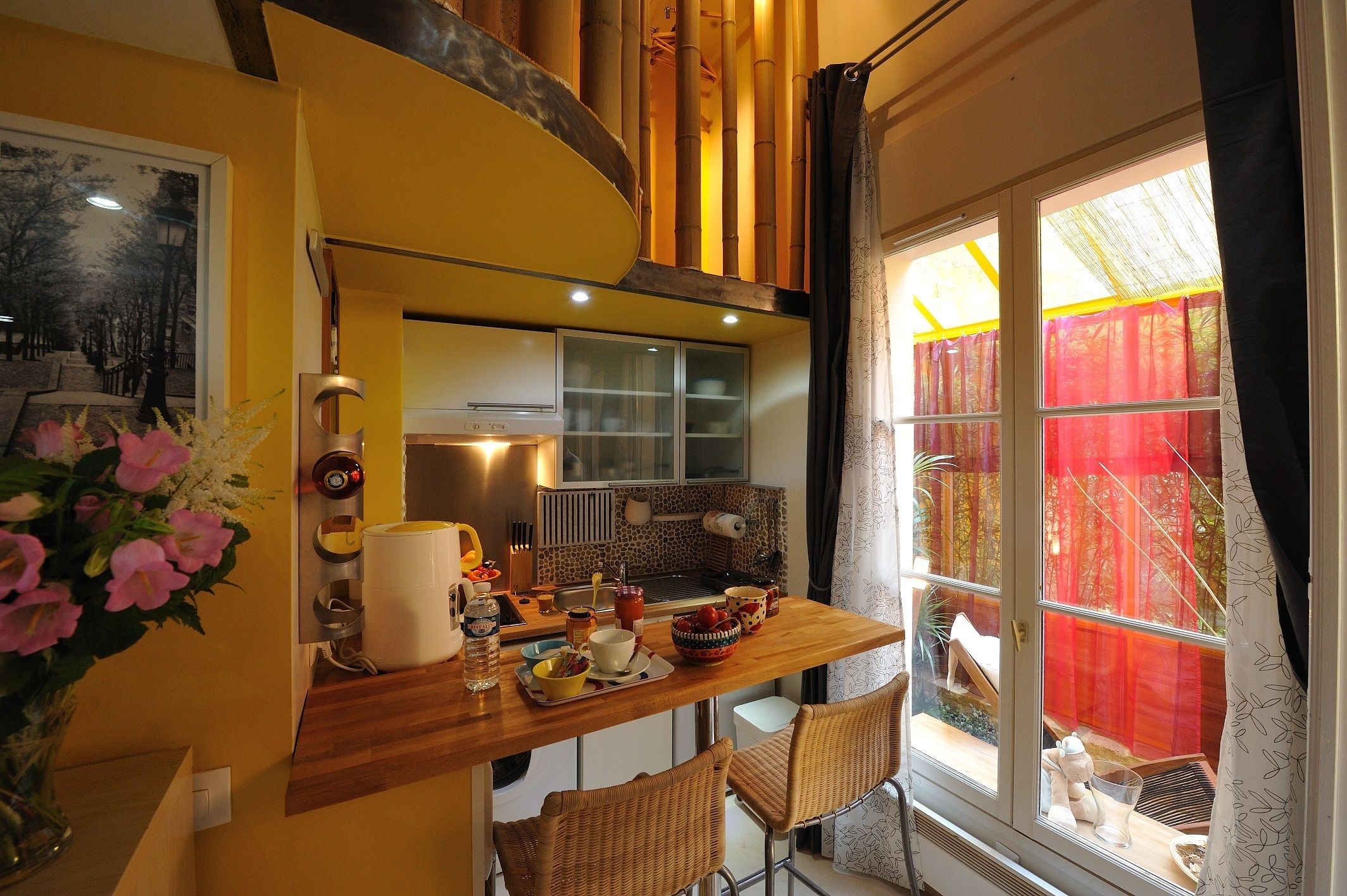 A cozy kitchen with a wooden countertop, a dining area with wicker chairs, and a large window with a view of a red fence and trees.