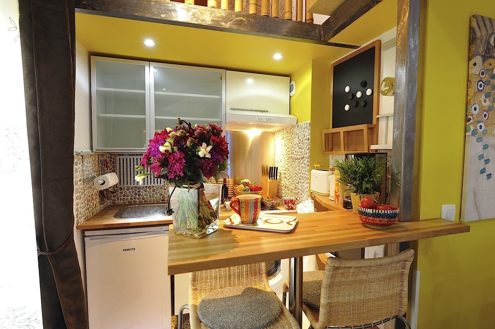 A compact kitchen with a wooden countertop, a small refrigerator, a window, and a vase of flowers.