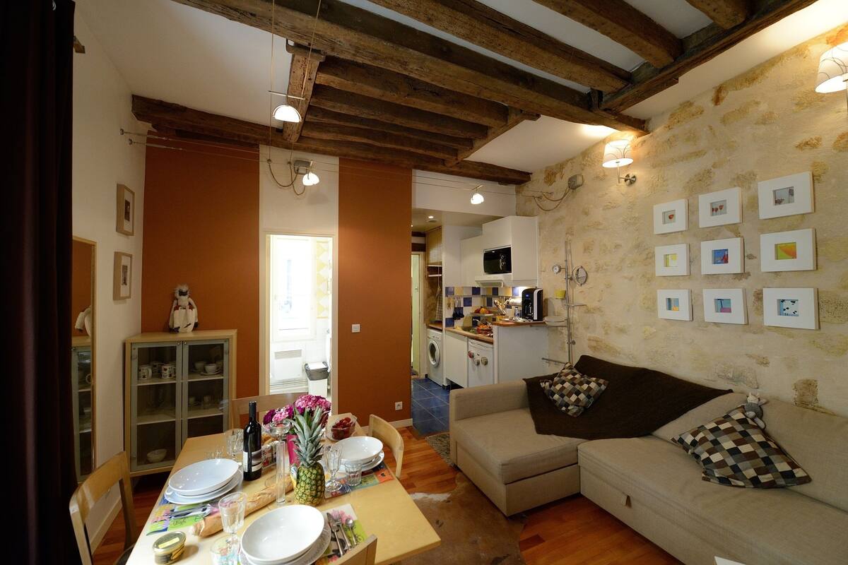 A cozy living room with a sofa, a dining table set with plates and a bottle, and a kitchen in the background.