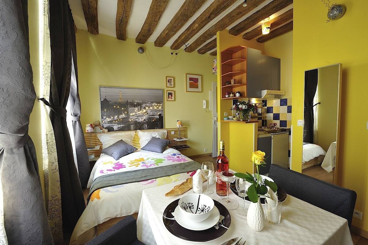 A hotel room with a bed, a dining table set with plates, glasses, and a bottle. There is a painting of a cityscape on the wall, and a kitchen area is visible in the background.