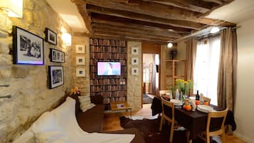 A cozy living room with a stone wall, wooden beams, a bookshelf, a TV, a dining area with a table set for two, and a sofa.