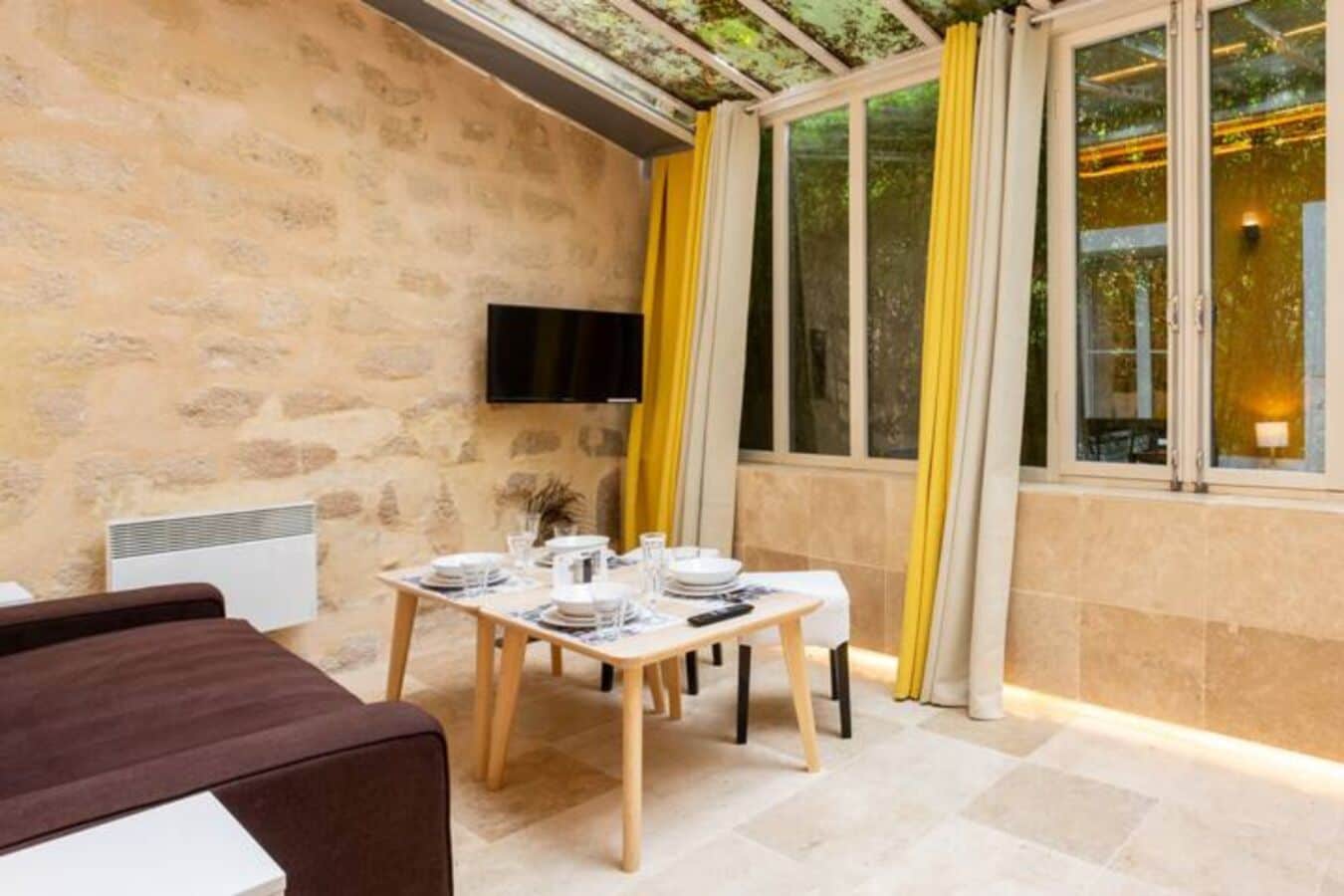 A dining area with a stone wall, a table set with plates and glasses, a flat-screen TV mounted on the wall, and yellow curtains on the windows.