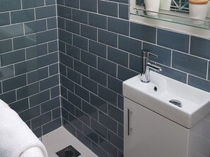 A bathroom with dark blue tiles, a white sink, and a glass shelf above the sink. - The Natterjack Inn (Shepton Mallet)