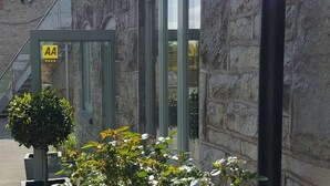 Un bâtiment en pierre avec une porte en verre portant l’inscription « AA » et un petit arbre au premier plan.
