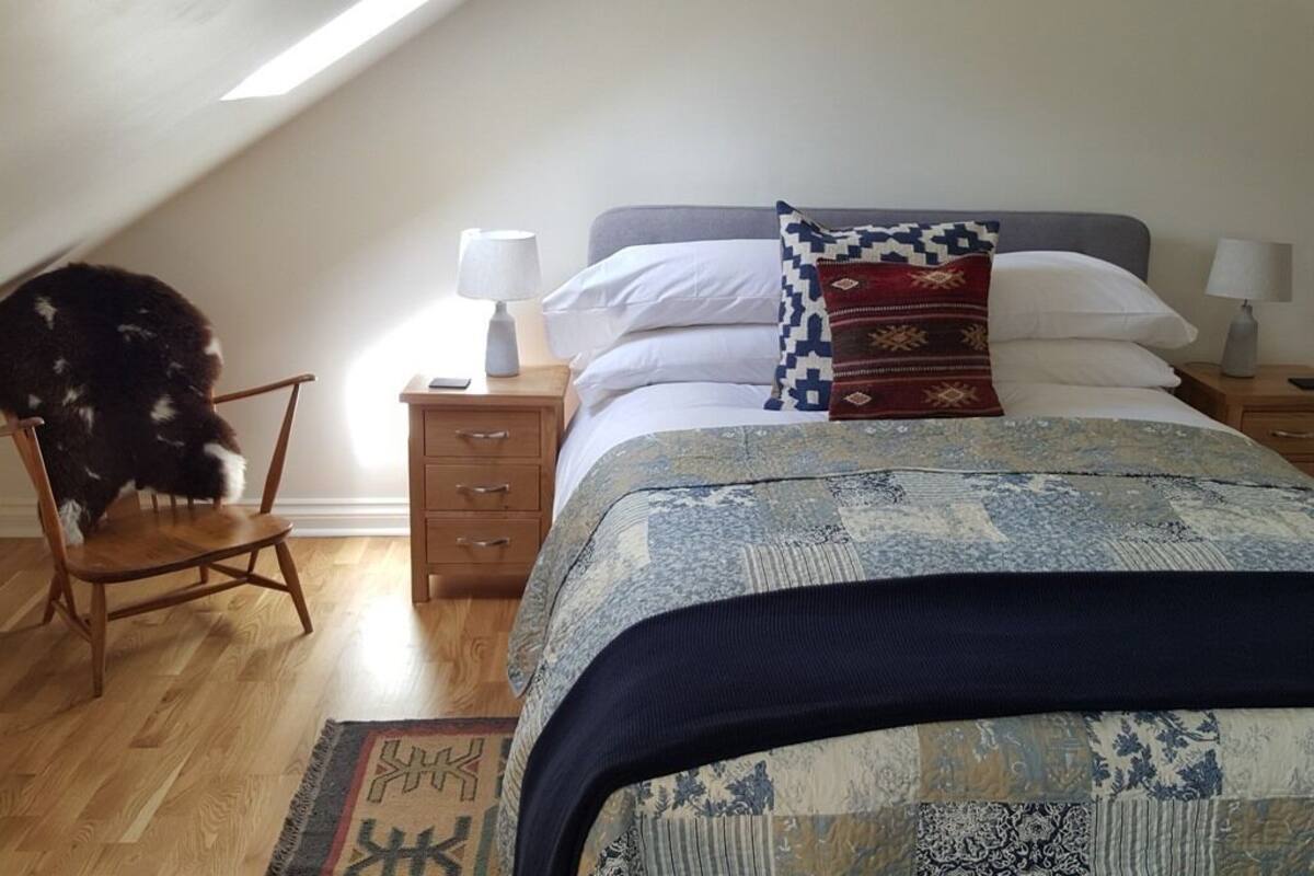 Une chambre avec un plafond en pente, un lit avec une literie à motifs, deux tables de chevet avec des lampes, une chaise en bois et un tapis sur le sol.
