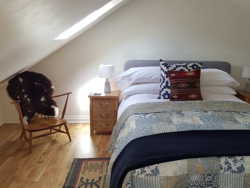 Et soveværelse med skrå vægge, en seng med mønstret sengetøj, en stol, et sengebord med en lampe og en lille måtte.